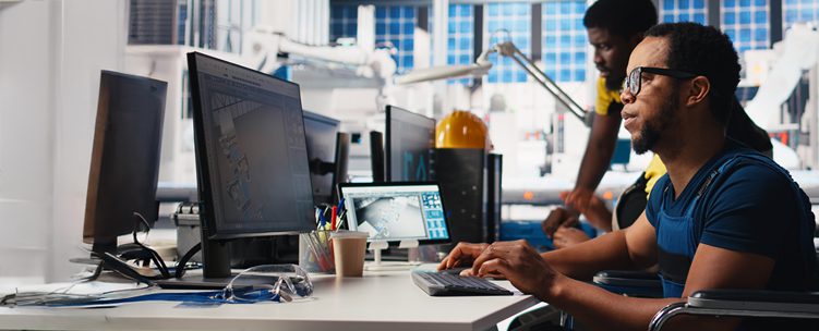 Man with glasses sitting at a computer in a manufacturing warehouse, looking at a screen with his hands on the keyboard.